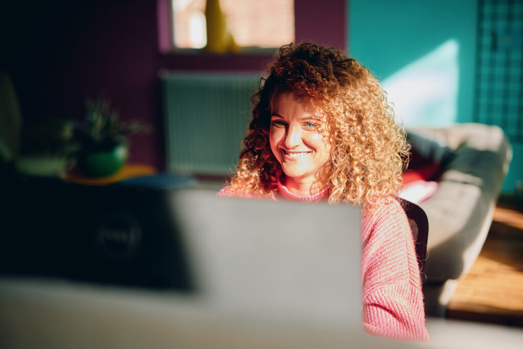 Vrouw met krullend haar en roze trui glimlacht terwijl ze achter een laptop werkt in een kleurrijke, zonnig kantoor.